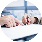 Close-up of two individuals at a desk, one pointing at a document while the other prepares to write, suggesting a review or signing of paperwork.