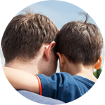 Two boys with their backs to the camera, one with an arm around the other, outdoors in daylight.