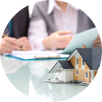 A small model house on a table with the blurred background of a person writing on a clipboard, suggesting a real estate, mortgage, or insurance context.